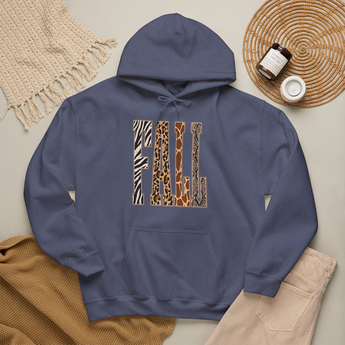 A Heather Navy pullover hoodie laid flat on a light beige surface, featuring large block letters across the chest filled with four animal print patterns: zebra, leopard, giraffe, and cheetah. The hoodie is styled with a tan knit blanket, beige pants, a woven placemat, and small jars in a cozy flat-lay arrangement.