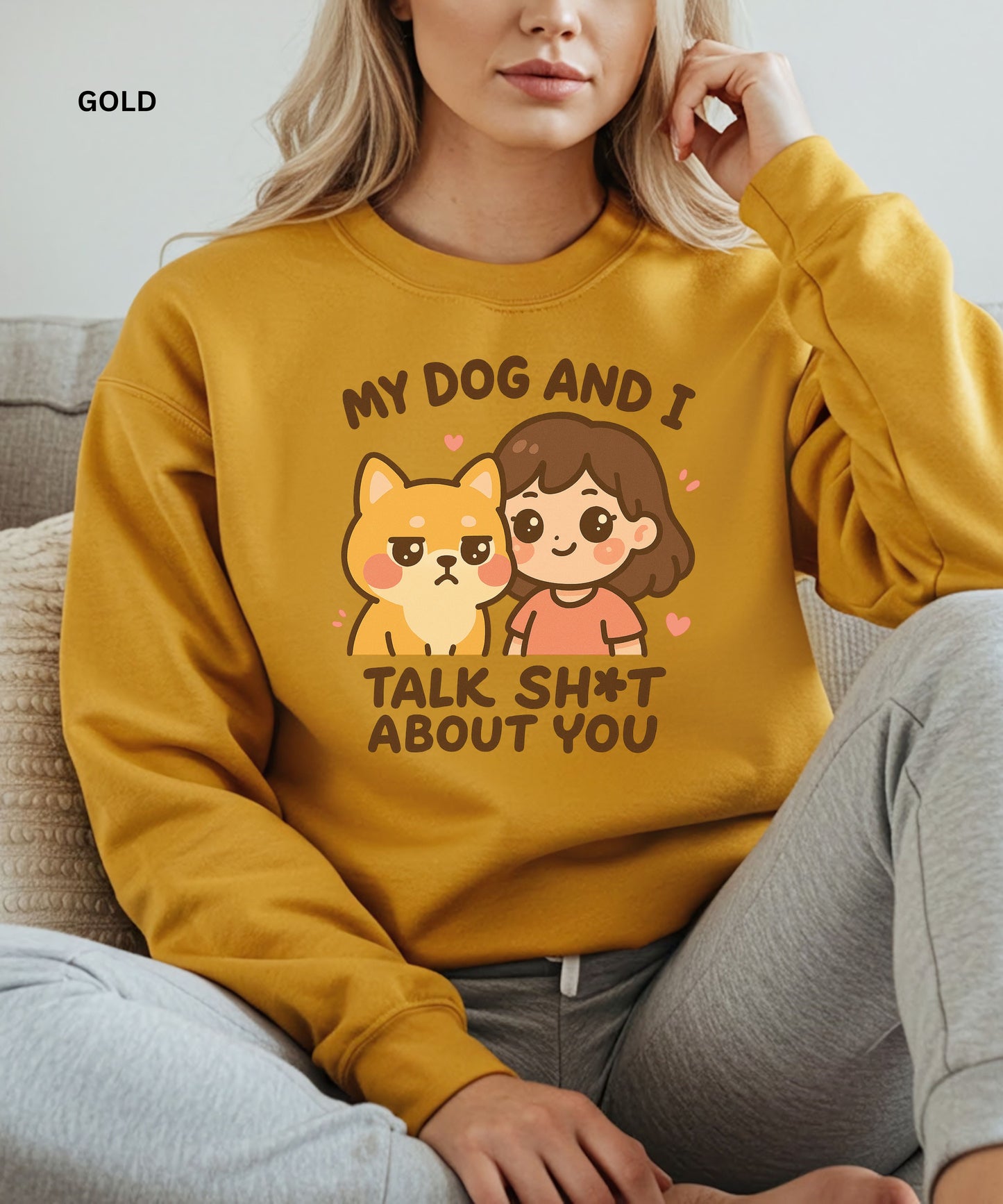 Person wearing a gold sweatshirt with a graphic and text that says"my dog and i talk shit about you", sitting on a couch.