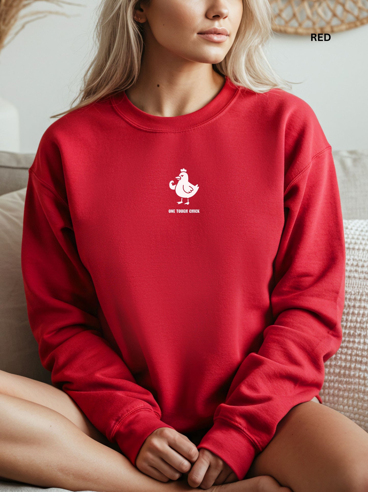 Person wearing a red sweatshirt with a black logo of a chicken and text " one tough chick", sitting on a textured surface.