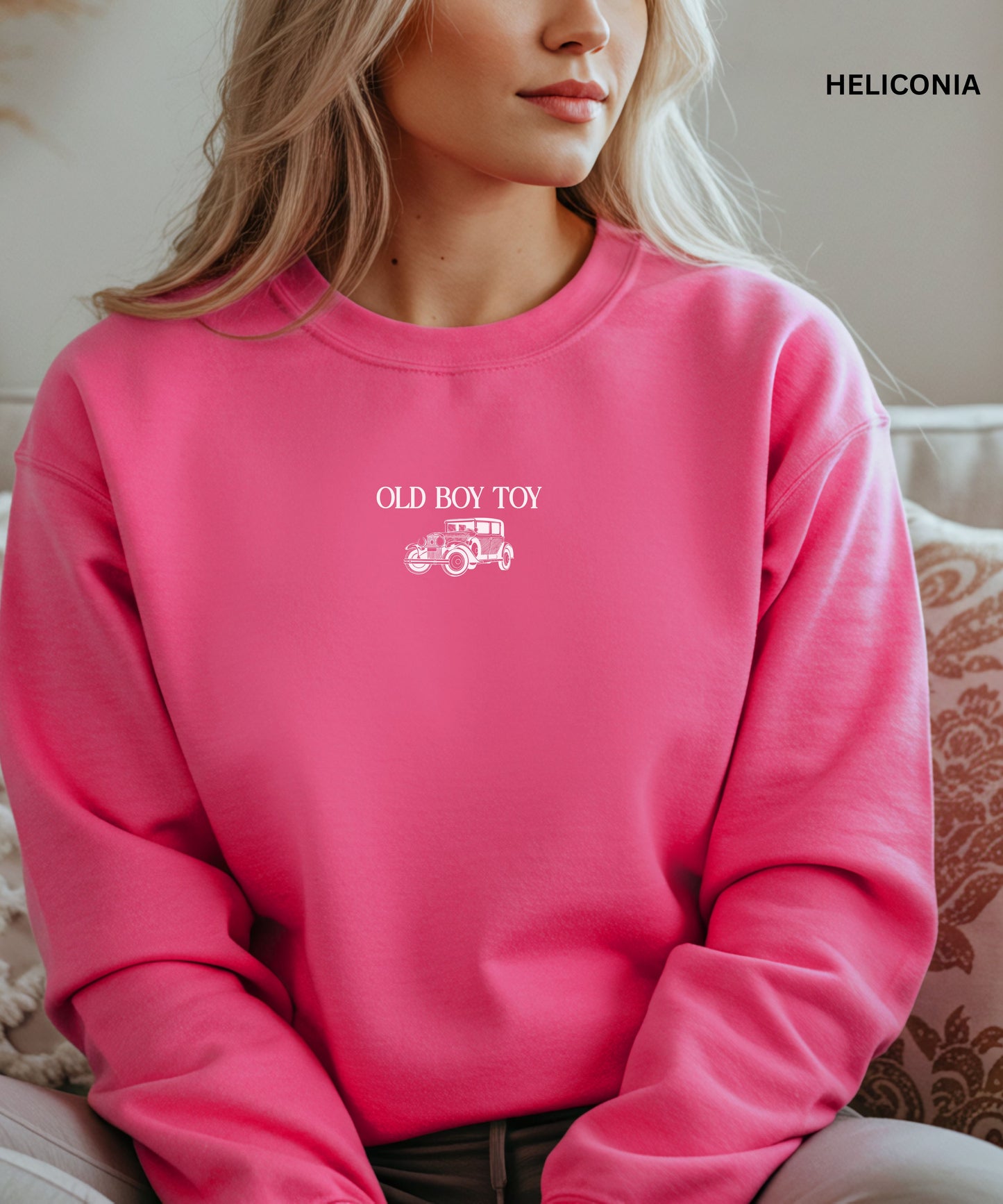 Person wearing a pink sweatshirt with 'Old Boy Toy' text and graphic, sitting on a patterned couch.