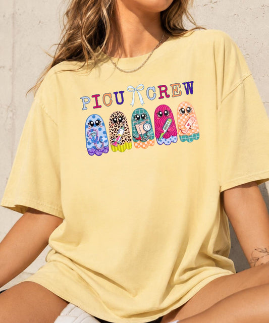Woman wearing butter yellow garment-dyed PICU Crew t-shirt with five colorful patterned ghosts holding nursing tools — stethoscope, syringe, blood pressure cuff, thermometer, and bandaids