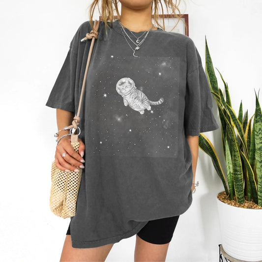 A woman wearing an oversized dark gray t-shirt with a white graphic of a cat in space, paired with black shorts and carrying a beige woven shoulder bag, standing next to a potted snake plant.