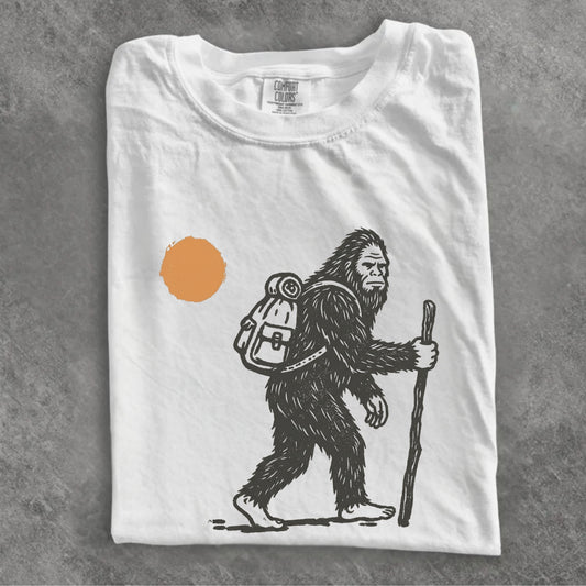 A folded white t-shirt with a black line art graphic of a Bigfoot hiker holding a staff, next to an orange sun, displayed on a textured gray background.