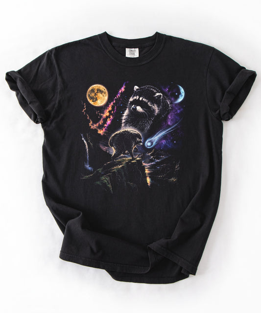 A black short-sleeve t-shirt with rolled cuffs, featuring a colorful graphic print of two raccoons in a cosmic space scene with a yellow moon and purple nebula.