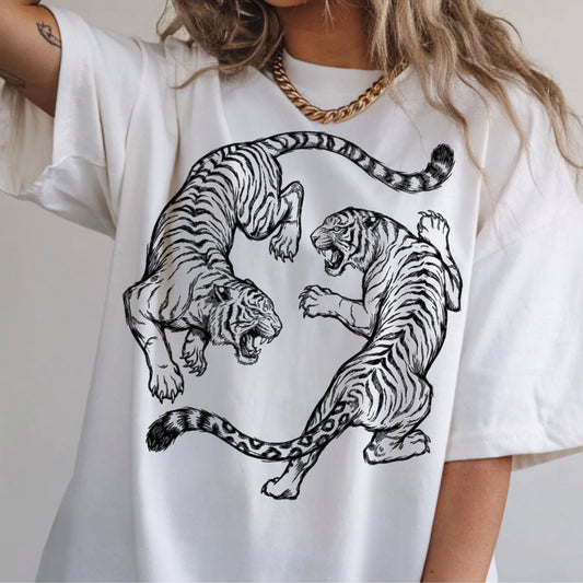 A person wearing an oversized white T-shirt with a large black line-art graphic of two roaring tigers facing each other. The photo is cropped from the neck to the hips, with a gold chain necklace visible against a light neutral background.