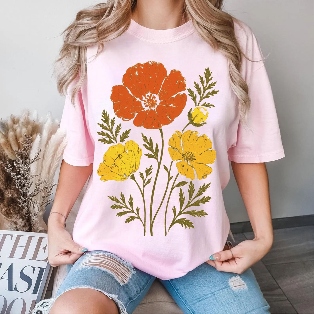 A woman wearing a Blossom pink short-sleeve t-shirt with a large graphic print of orange and yellow poppy flowers with green leaves, paired with distressed denim shorts.