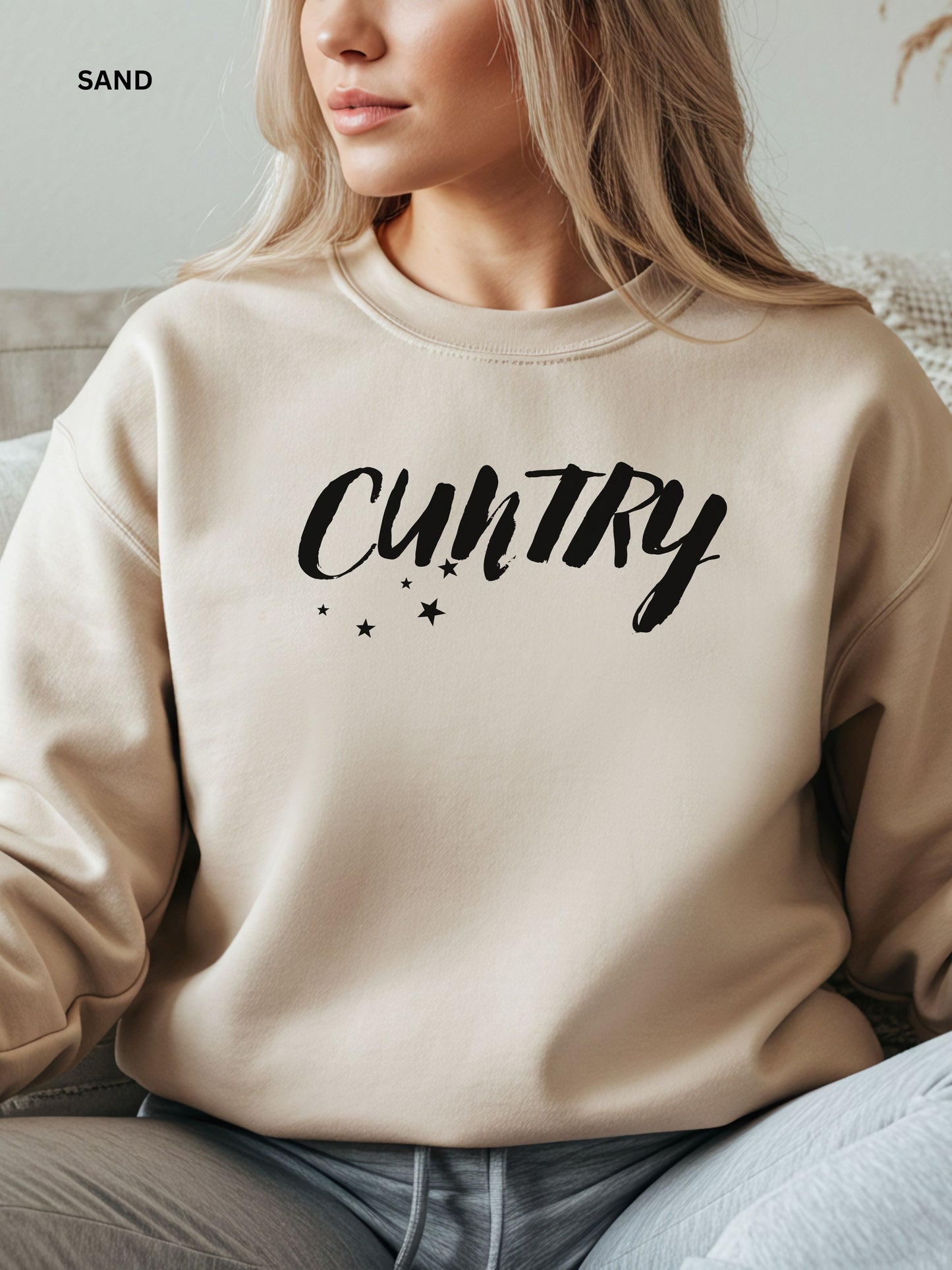 Person wearing a sand sweatshirt with 'CUNTRY' printed on it, sitting on a couch.