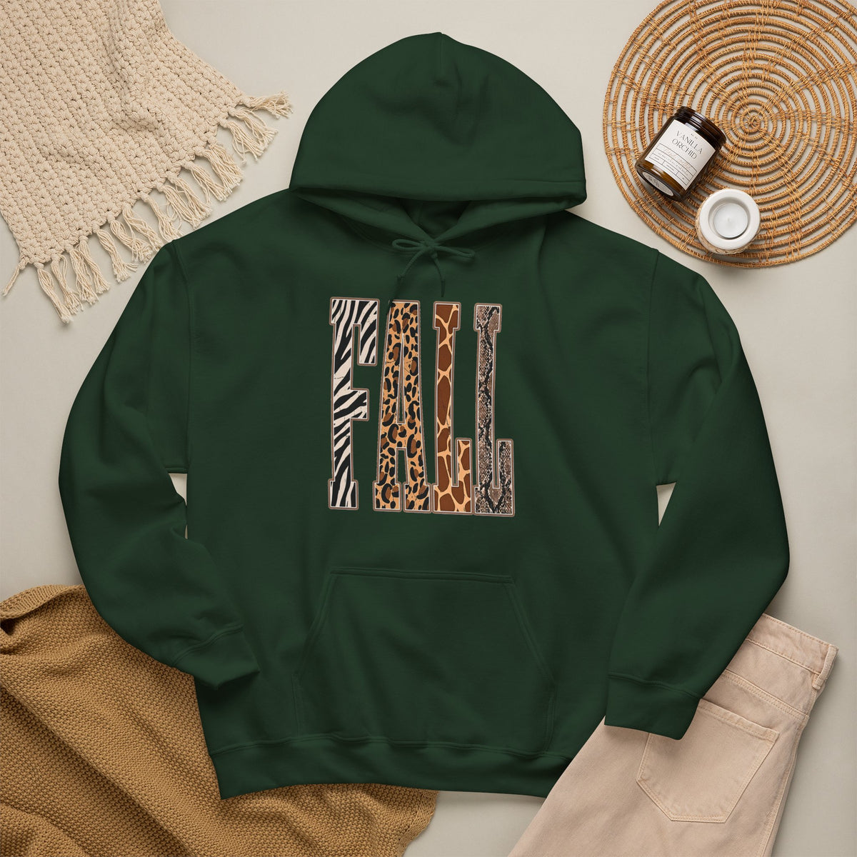 A forest green pullover hoodie laid flat on a light beige surface, featuring large block letters across the chest filled with four animal print patterns: zebra, leopard, giraffe, and cheetah. The hoodie is styled with a tan knit blanket, beige pants, a woven placemat, and small jars in a cozy flat-lay arrangement.