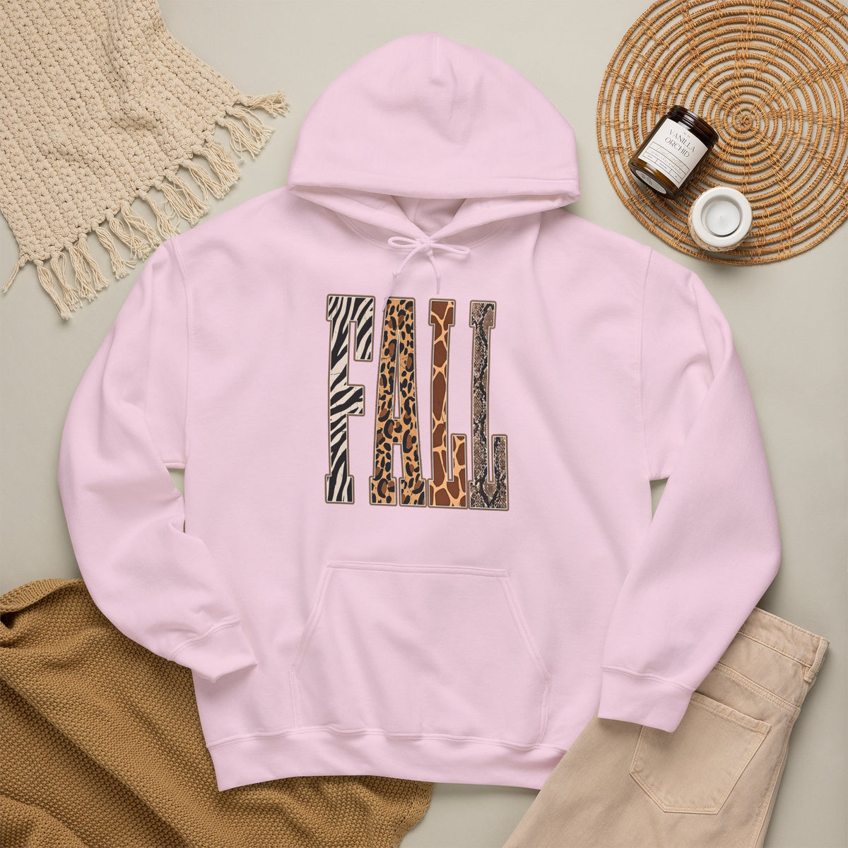 A Light pink pullover hoodie laid flat on a light beige surface, featuring large block letters across the chest filled with four animal print patterns: zebra, leopard, giraffe, and cheetah. The hoodie is styled with a tan knit blanket, beige pants, a woven placemat, and small jars in a cozy flat-lay arrangement.