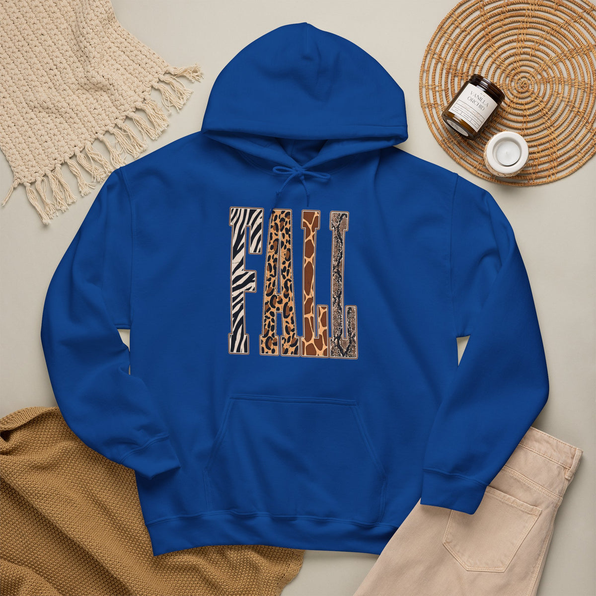 A Royal blue pullover hoodie laid flat on a light beige surface, featuring large block letters across the chest filled with four animal print patterns: zebra, leopard, giraffe, and cheetah. The hoodie is styled with a tan knit blanket, beige pants, a woven placemat, and small jars in a cozy flat-lay arrangement.