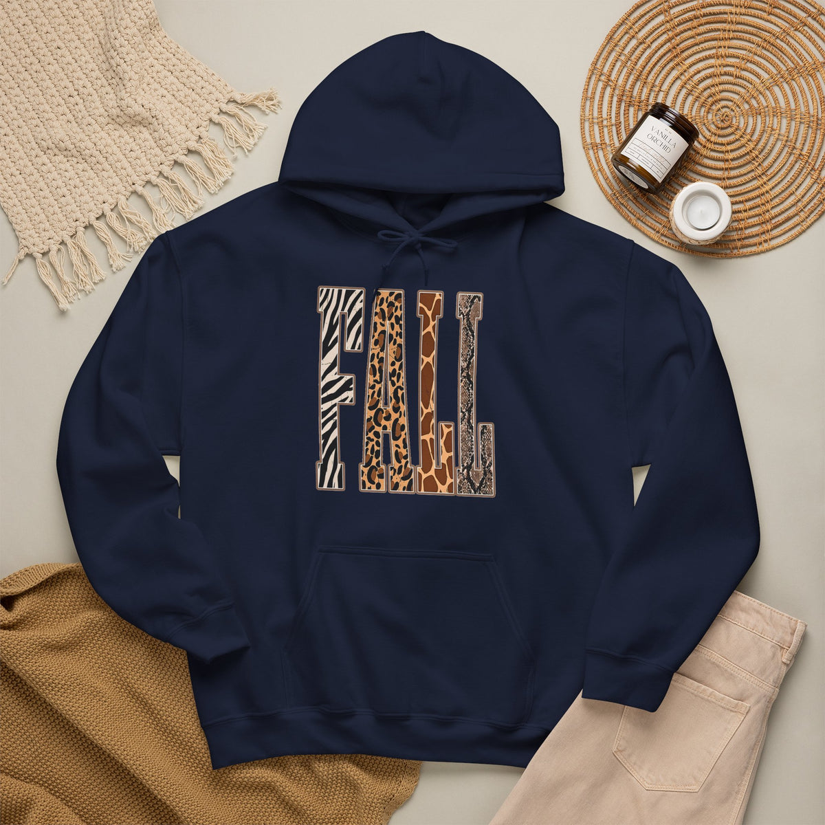 A navy pullover hoodie laid flat on a light beige surface, featuring large block letters across the chest filled with four animal print patterns: zebra, leopard, giraffe, and cheetah. The hoodie is styled with a tan knit blanket, beige pants, a woven placemat, and small jars in a cozy flat-lay arrangement.