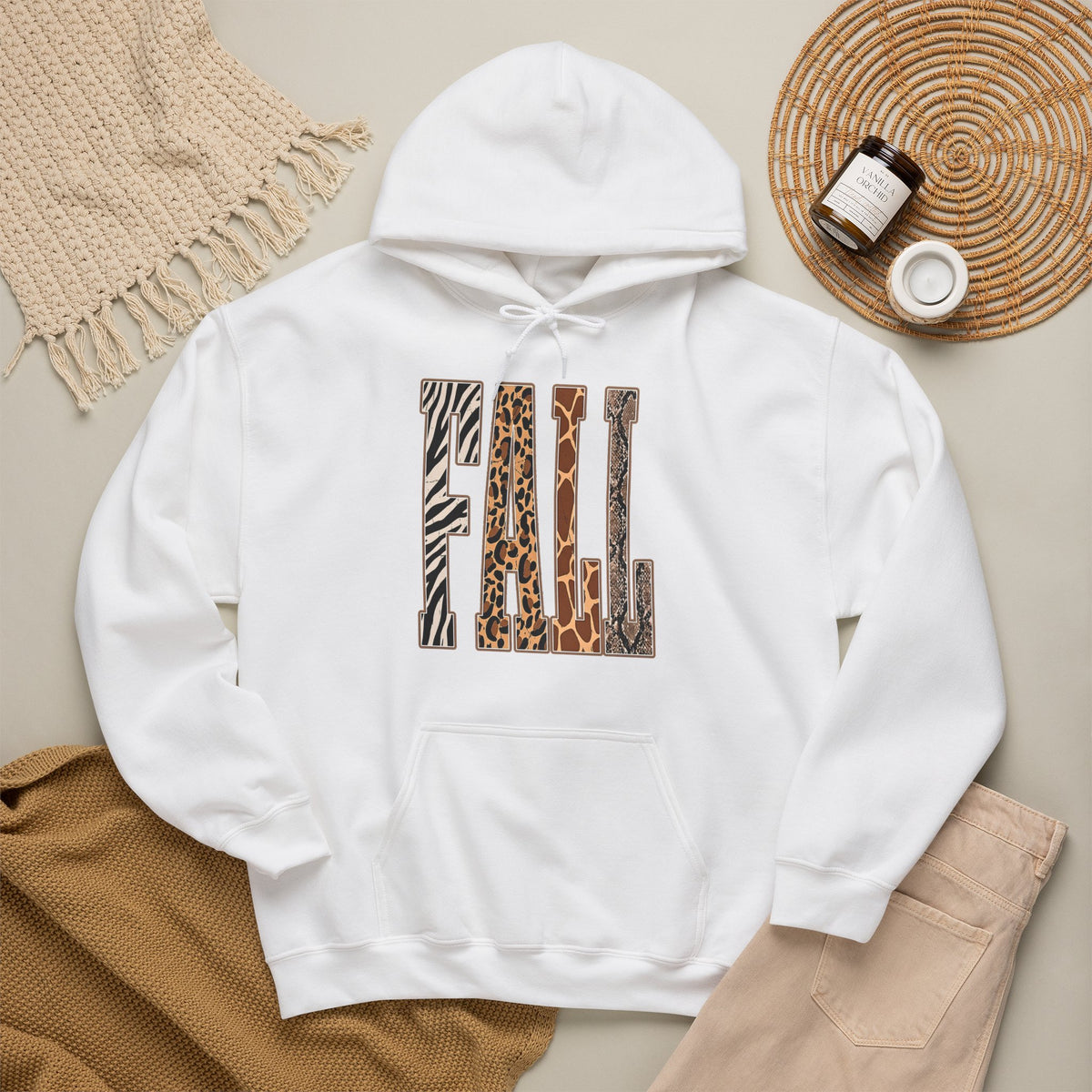 A white pullover hoodie laid flat on a light beige surface, featuring large block letters across the chest filled with four animal print patterns: zebra, leopard, giraffe, and cheetah. The hoodie is styled with a tan knit blanket, beige pants, a woven placemat, and small jars in a cozy flat-lay arrangement.