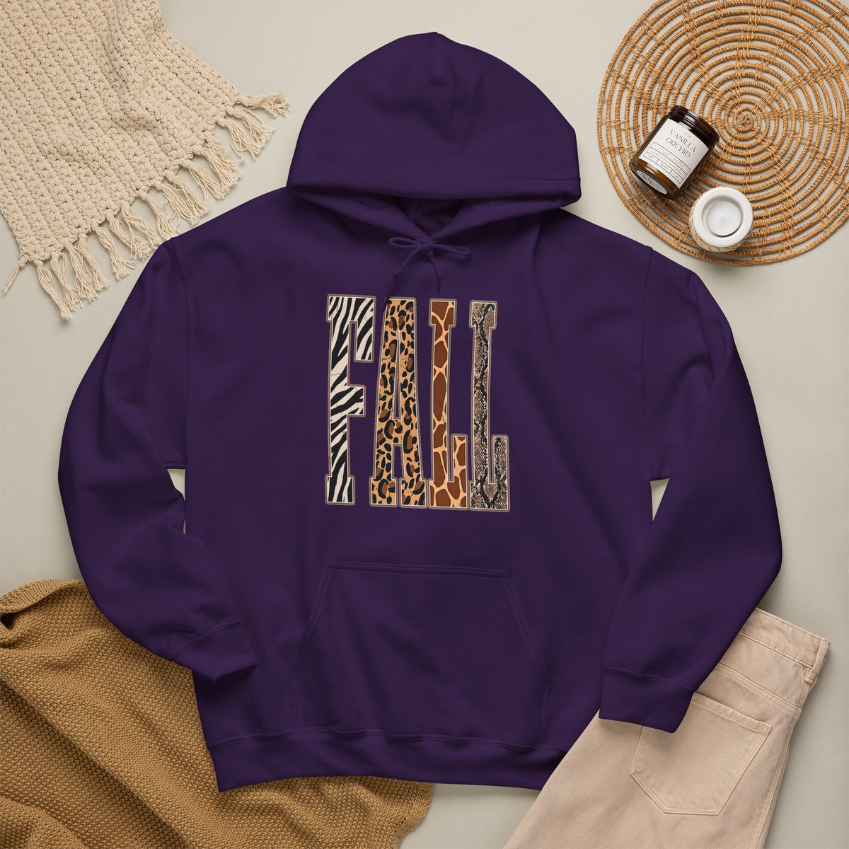 A Purple pullover hoodie laid flat on a light beige surface, featuring large block letters across the chest filled with four animal print patterns: zebra, leopard, giraffe, and cheetah. The hoodie is styled with a tan knit blanket, beige pants, a woven placemat, and small jars in a cozy flat-lay arrangement.
