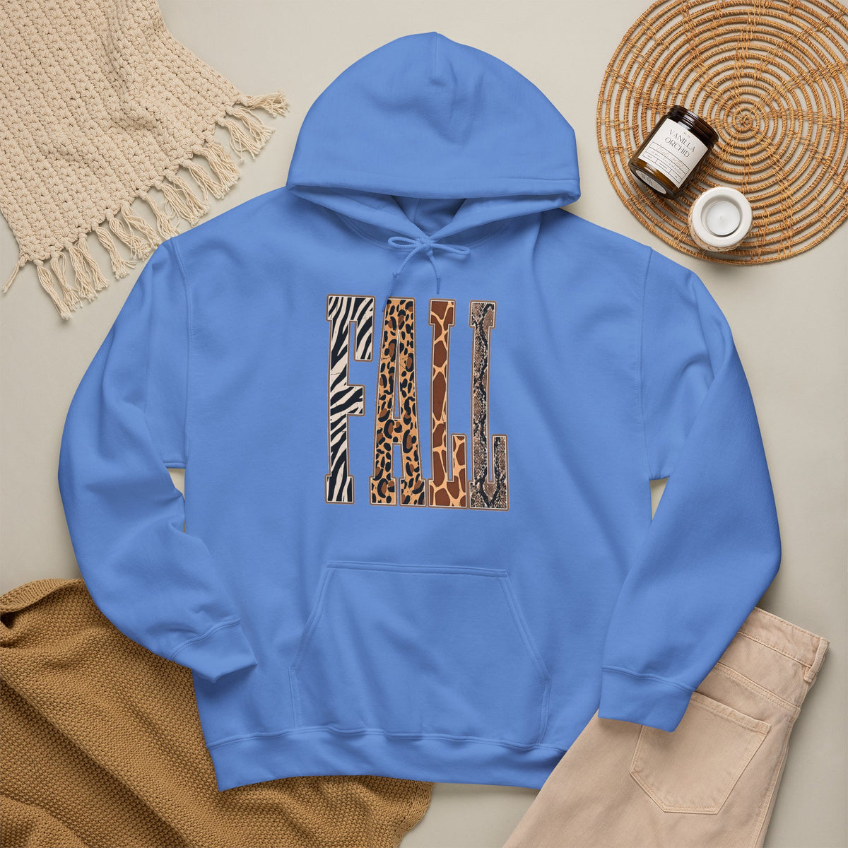 A Carolina Blue pullover hoodie laid flat on a light beige surface, featuring large block letters across the chest filled with four animal print patterns: zebra, leopard, giraffe, and cheetah. The hoodie is styled with a tan knit blanket, beige pants, a woven placemat, and small jars in a cozy flat-lay arrangement.
