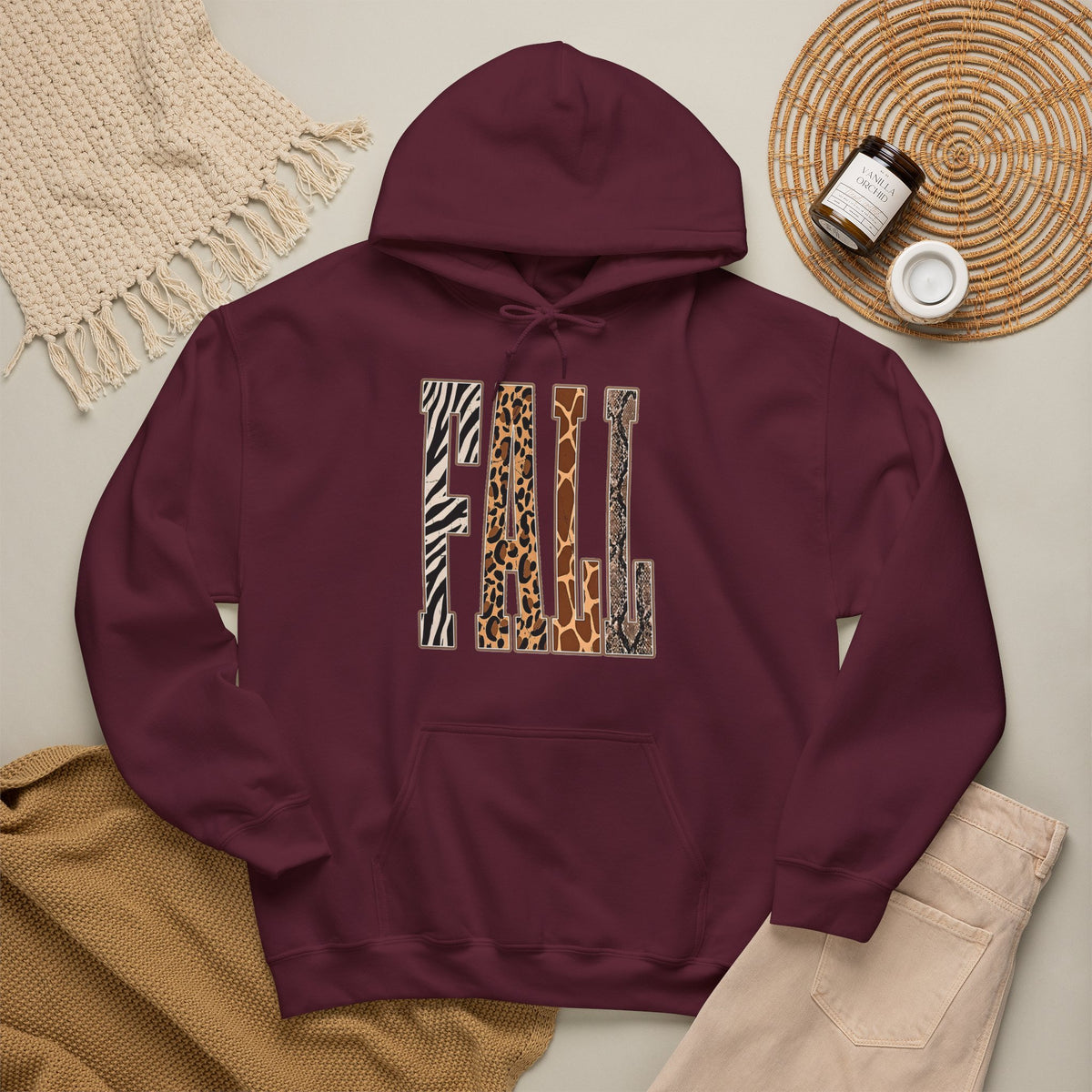  A maroon pullover hoodie laid flat on a light beige surface, featuring large block letters across the chest filled with four animal print patterns: zebra, leopard, giraffe, and cheetah. The hoodie is styled with a tan knit blanket, beige pants, a woven placemat, and small jars in a cozy flat-lay arrangement.