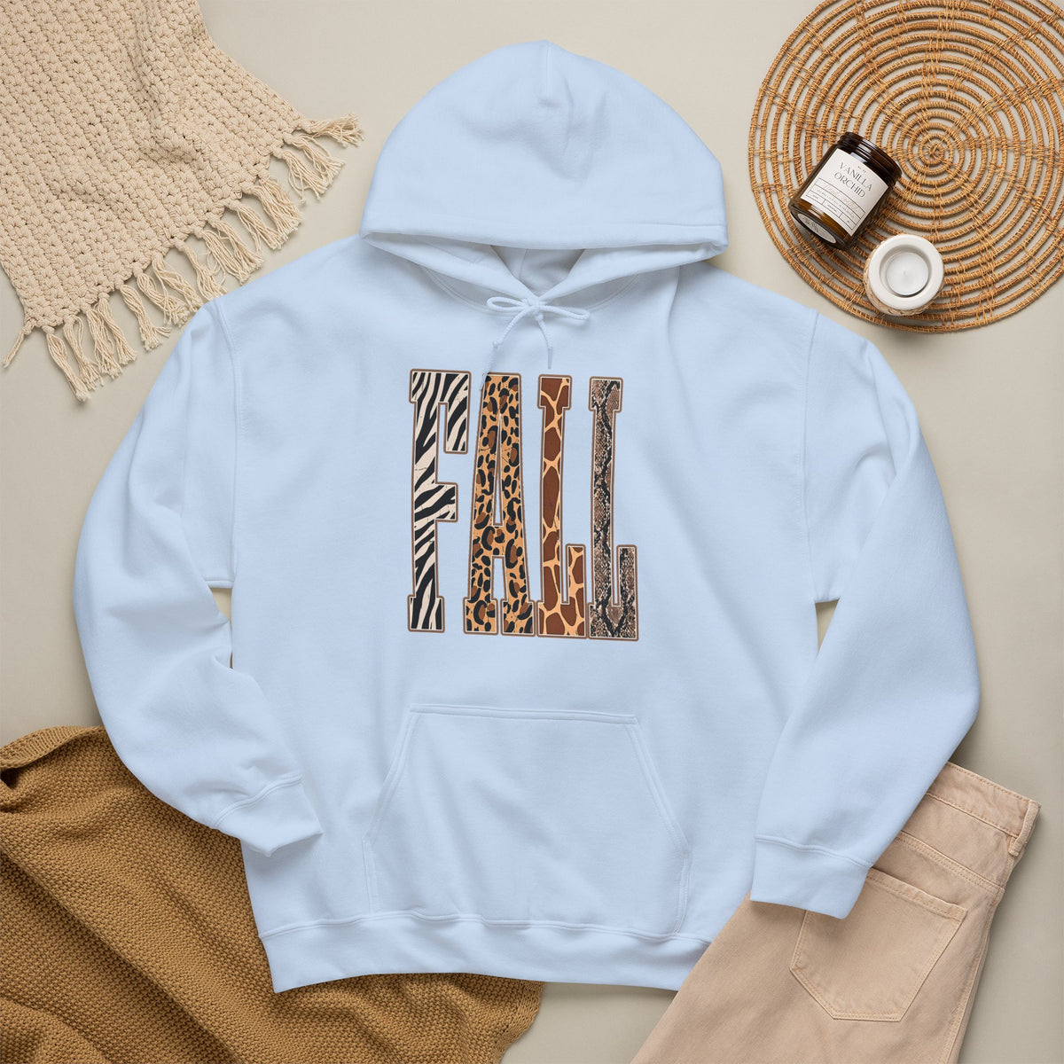 A Light Blue pullover hoodie laid flat on a light beige surface, featuring large block letters across the chest filled with four animal print patterns: zebra, leopard, giraffe, and cheetah. The hoodie is styled with a tan knit blanket, beige pants, a woven placemat, and small jars in a cozy flat-lay arrangement.