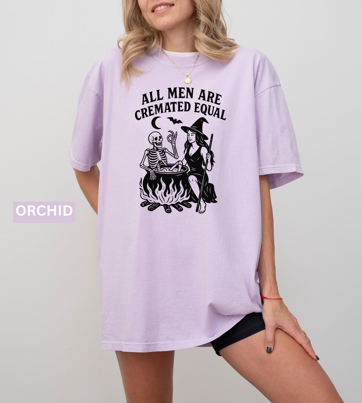 Person wearing an orchid graphic t-shirt with a skeleton and witch design, sitting on the ground.