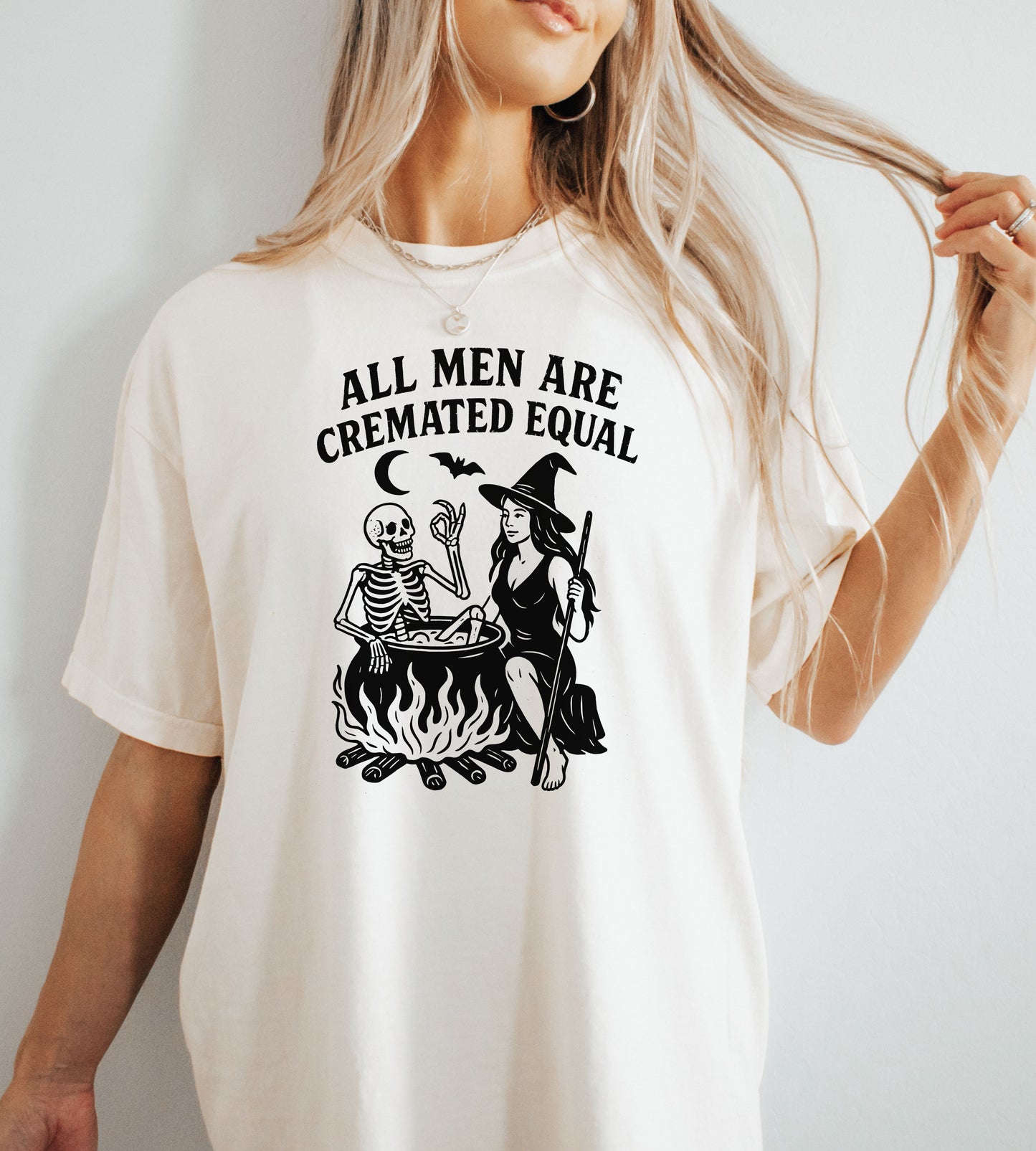 Person wearing an ivory graphic t-shirt with a skeleton and witch design, sitting on the ground.