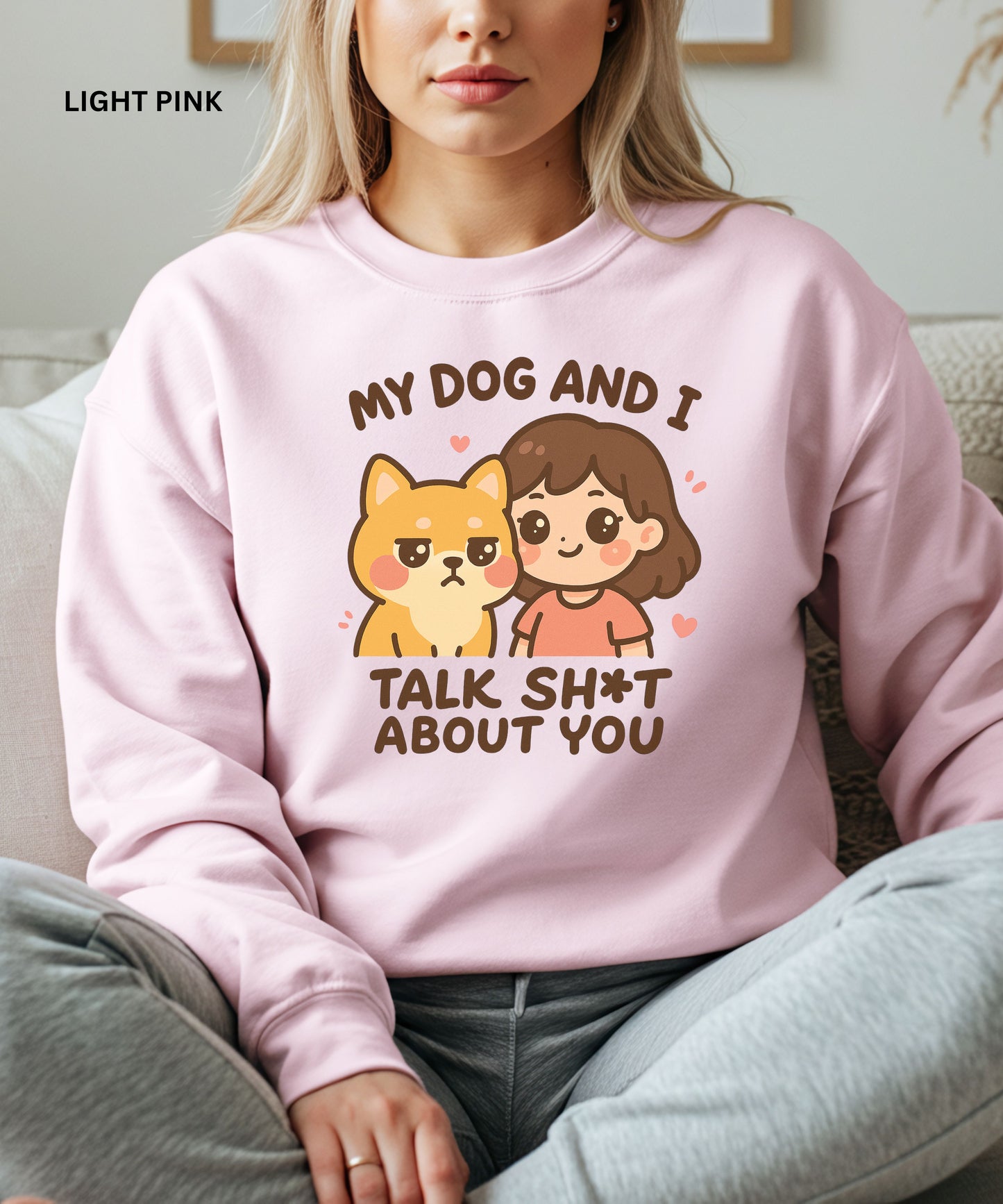 Person wearing a light pink sweatshirt with a graphic and text that says"my dog and i talk shit about you", sitting on a couch.