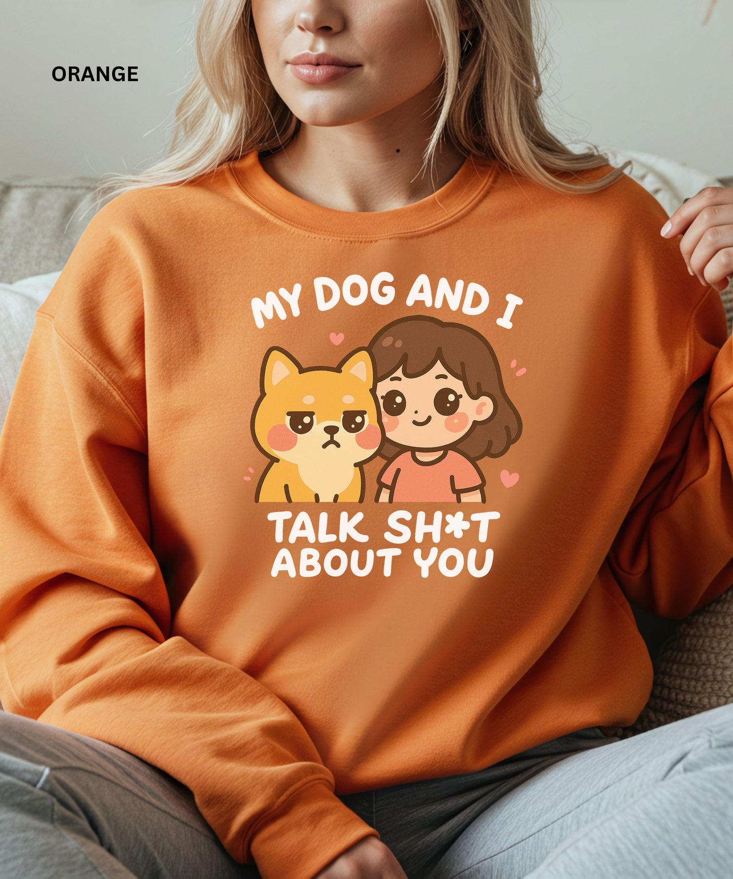 Person wearing an orange sweatshirt with a graphic and text that says"my dog and i talk shit about you", sitting on a couch.