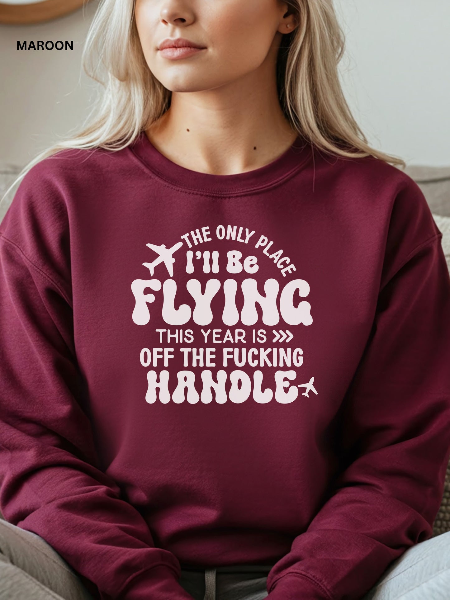Person wearing a maroon sweatshirt with a humorous text design that says "the only place ill be flying is off the fucking handle, sitting on a couch.