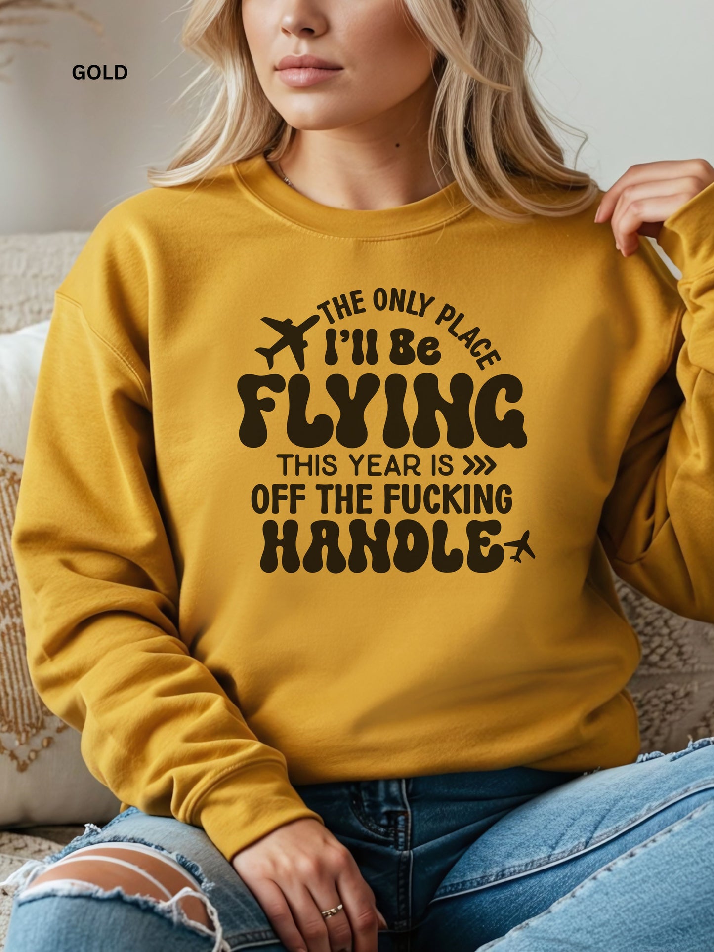 Person wearing a gold sweatshirt with a humorous text design that says "the only place ill be flying is off the fucking handle, sitting on a couch.