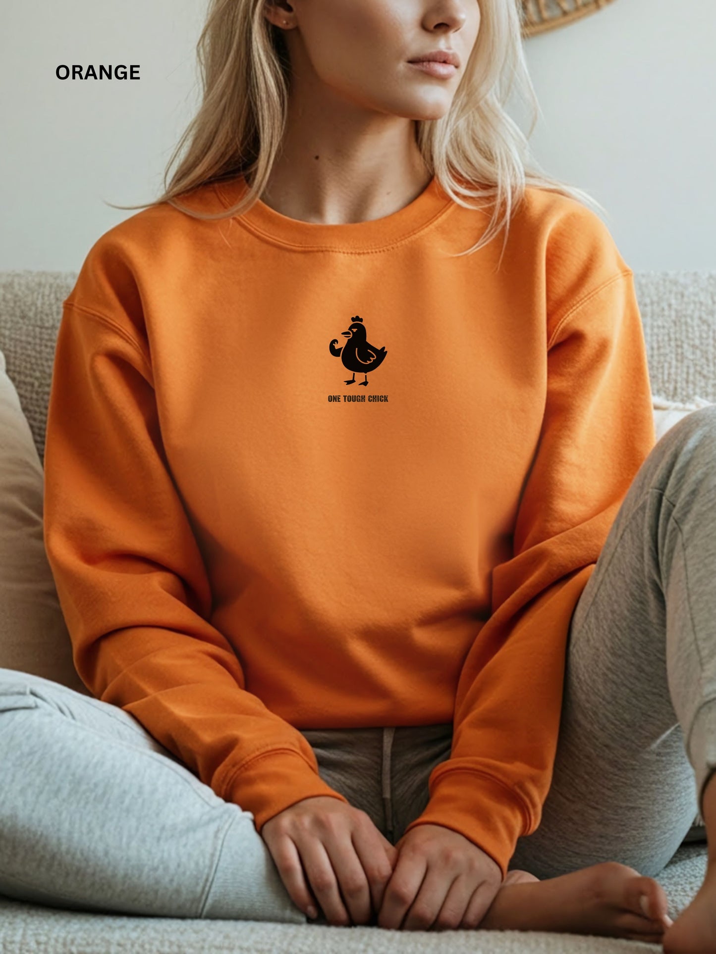 Person wearing a orange sweatshirt with a black logo of a chicken and text " one tough chick", sitting on a textured surface.