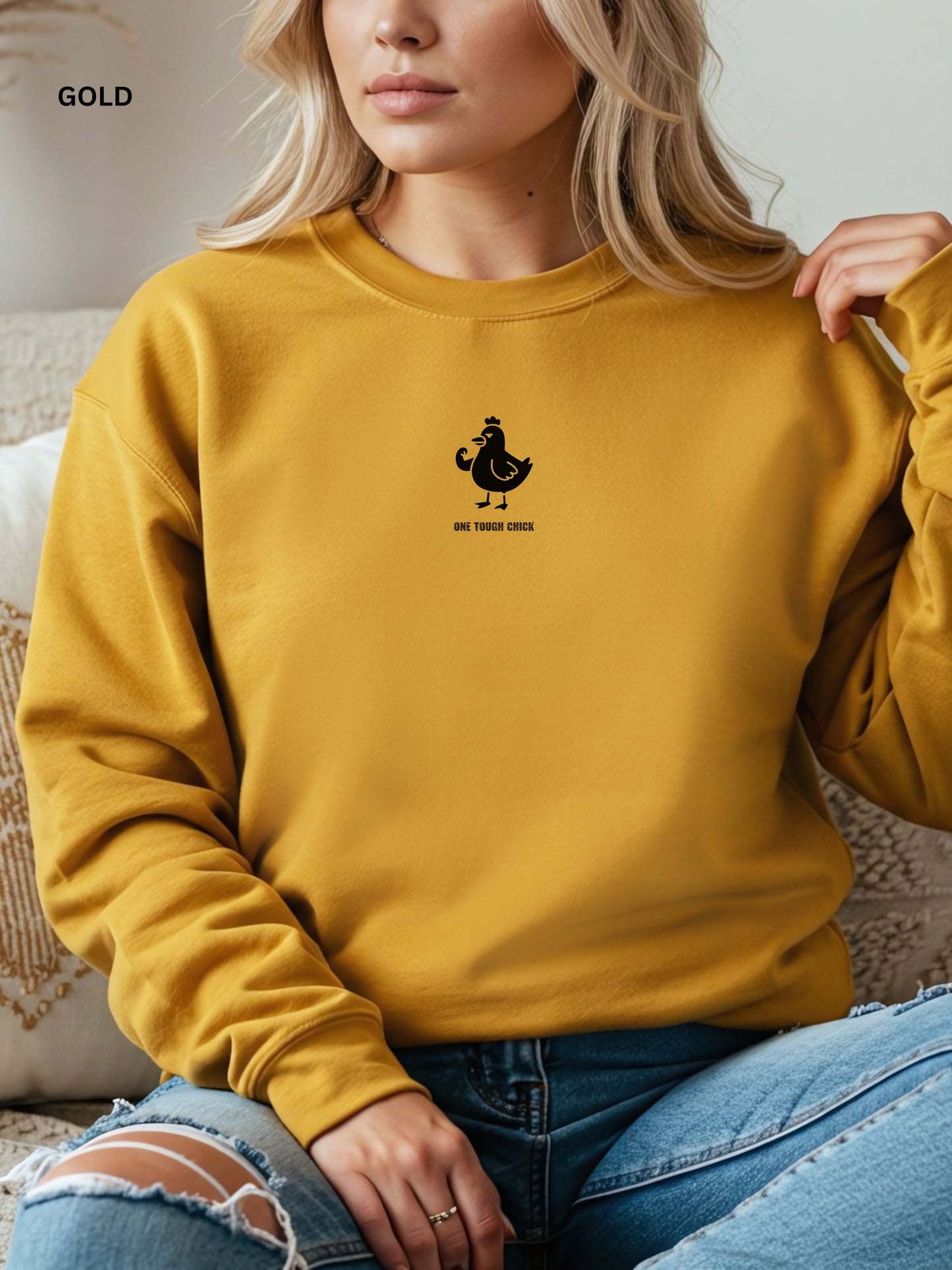 Person wearing a gold sweatshirt with a black logo of a chicken and text " one tough chick", sitting on a textured surface.