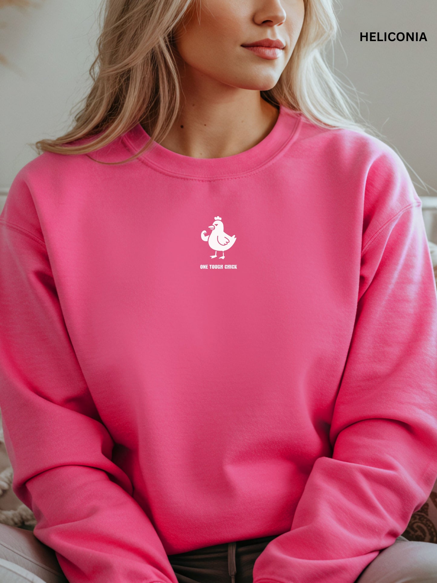 Person wearing a heliconia sweatshirt with a black logo of a chicken and text " one tough chick", sitting on a textured surface.