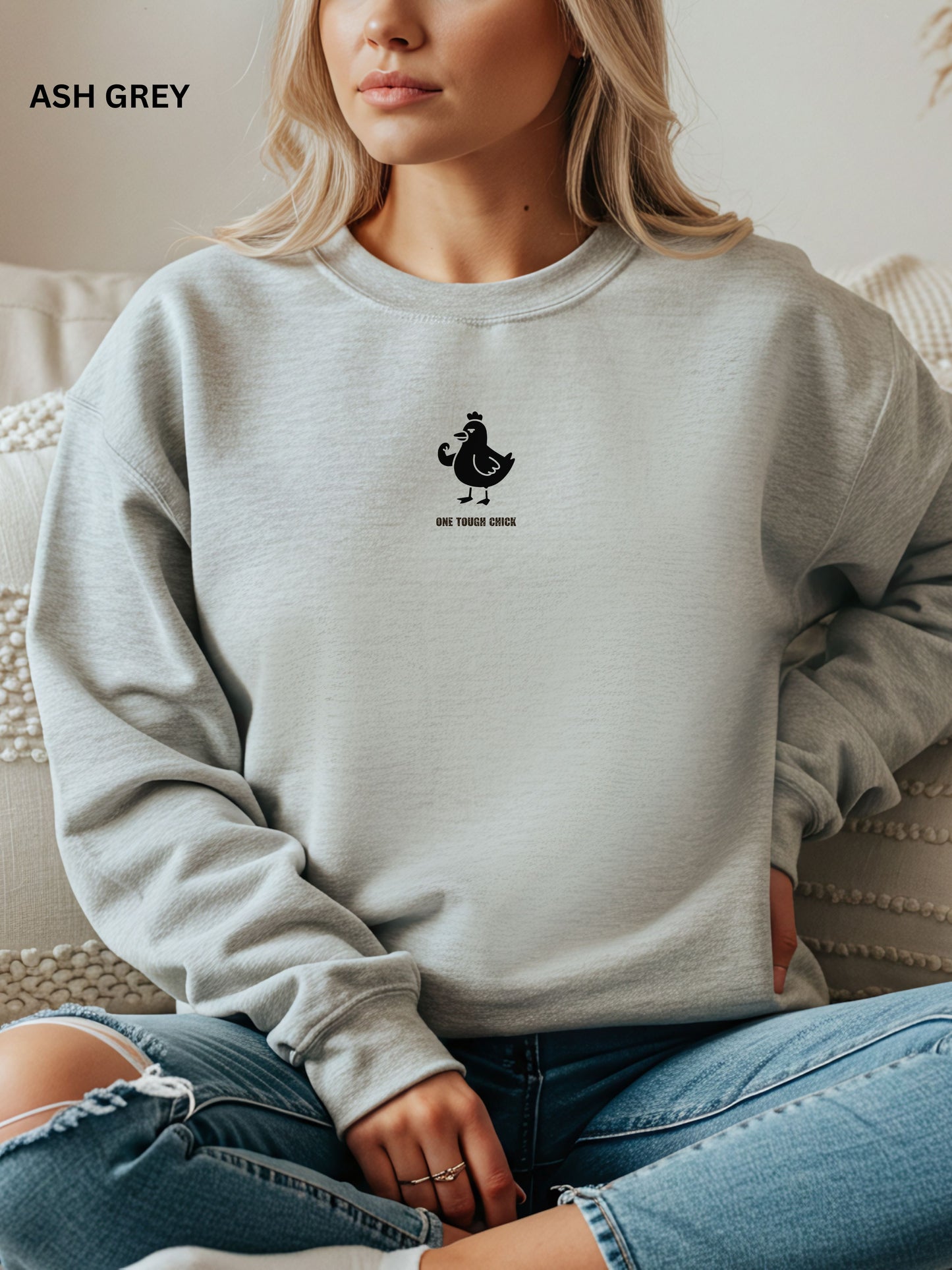 Person wearing an ash grey sweatshirt with a black logo of a chicken and text " one tough chick", sitting on a textured surface.