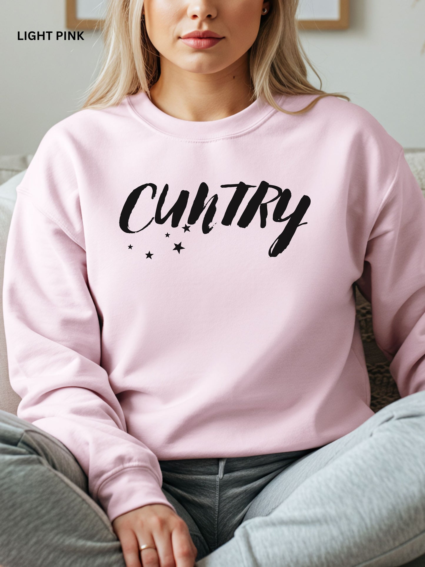 Person wearing a light pink  sweatshirt with 'CUNTRY' printed on it, sitting on a couch.