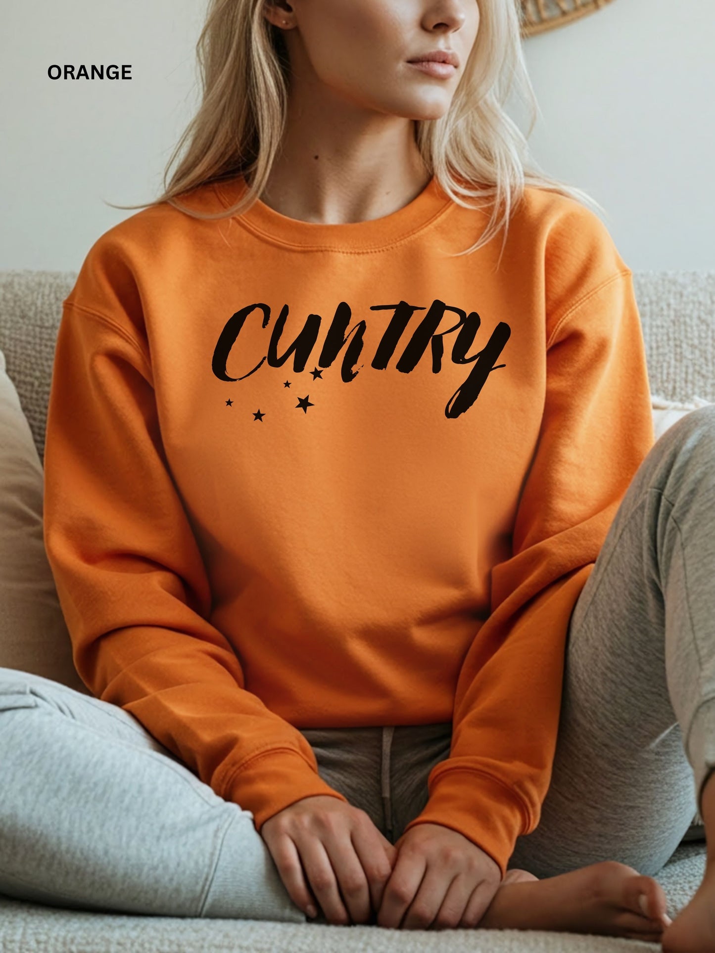 Person wearing an orange  sweatshirt with 'CUNTRY' printed on it, sitting on a couch.