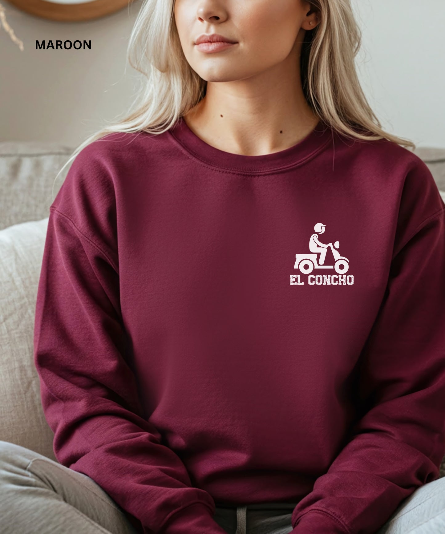 Person wearing a maroon sweatshirt with a logo and text that says" el concho", sitting on a couch.