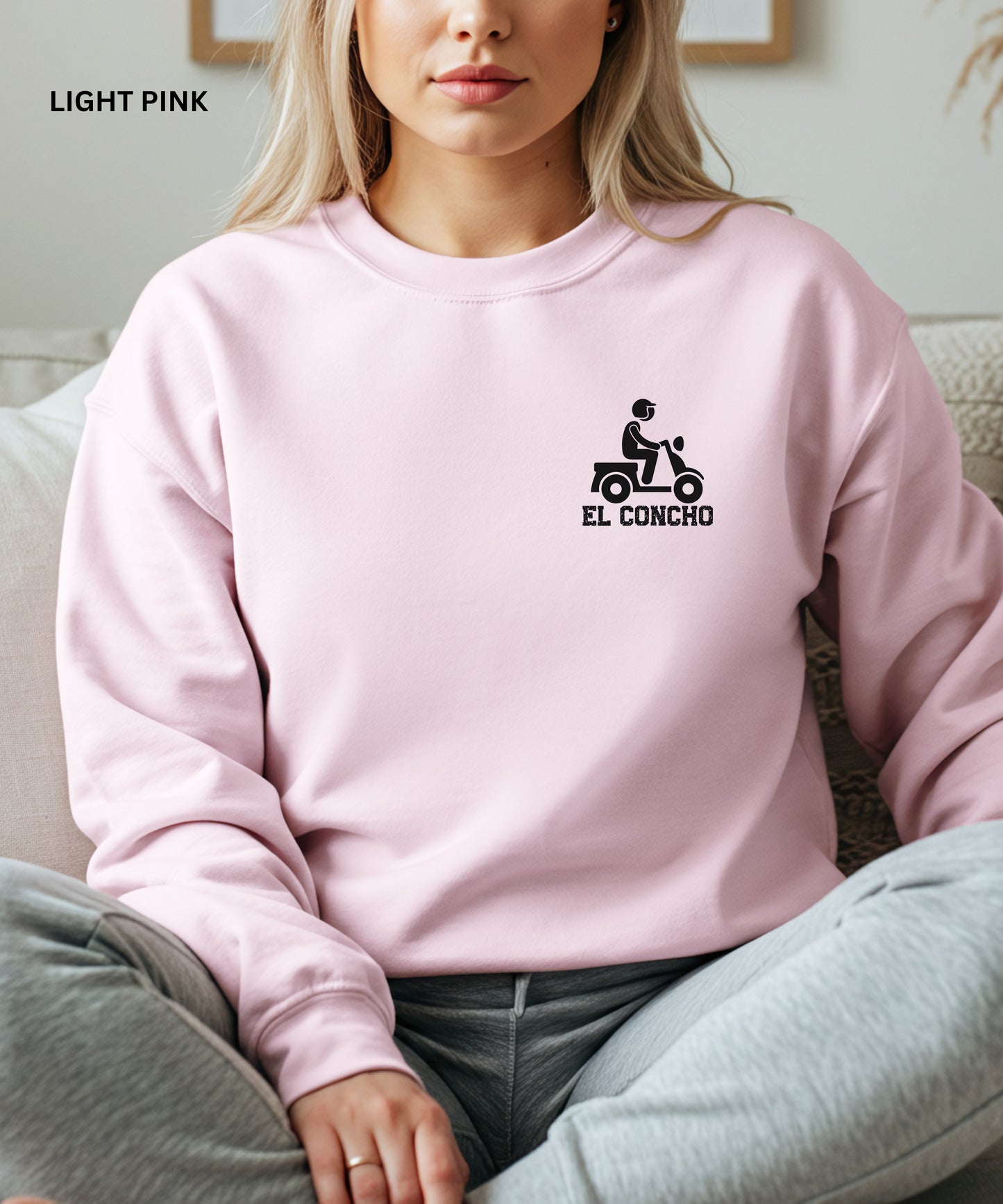 Person wearing a  light pink sweatshirt with a logo and text that says" el concho", sitting on a couch.