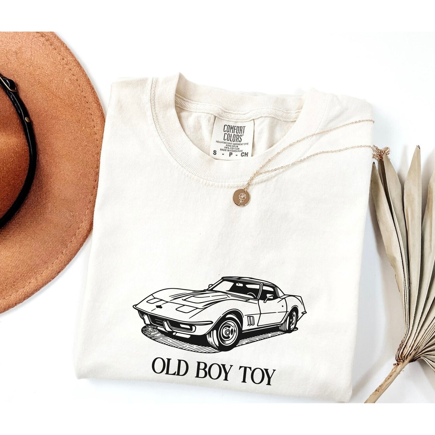 White t-shirt with car graphic and 'Old Boy Toy' text, placed on a white surface with a hat and dried plant.