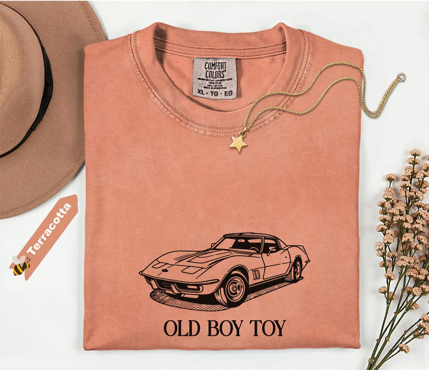 Terracotta-colored t-shirt with car graphic and 'Old Boy Toy' text, placed on a white surface with a hat and flowers.
