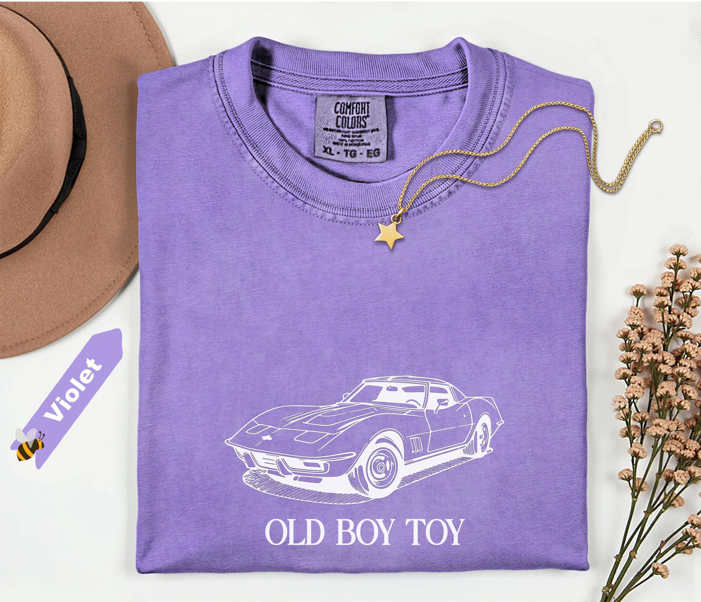 Purple t-shirt with car graphic and 'Old Boy Toy' text, placed on a white surface with a hat and flowers.