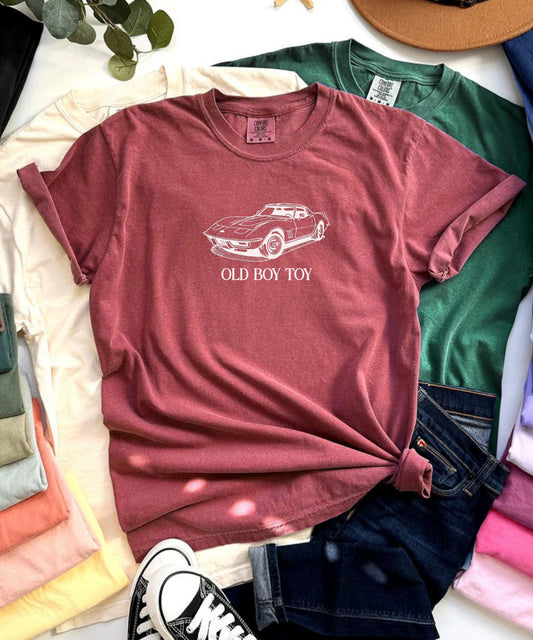 Maroon t-shirt with 'Old Boy Toy' graphic on a bed with colorful clothes and a hat.