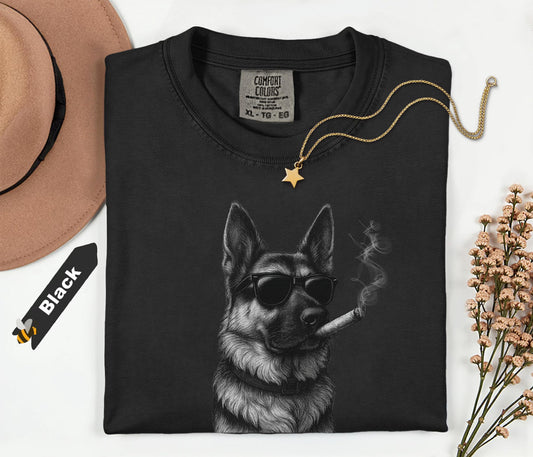 Black t-shirt with a graphic of a dog smoking a cigarette, placed on a white surface with a hat and dried flowers.