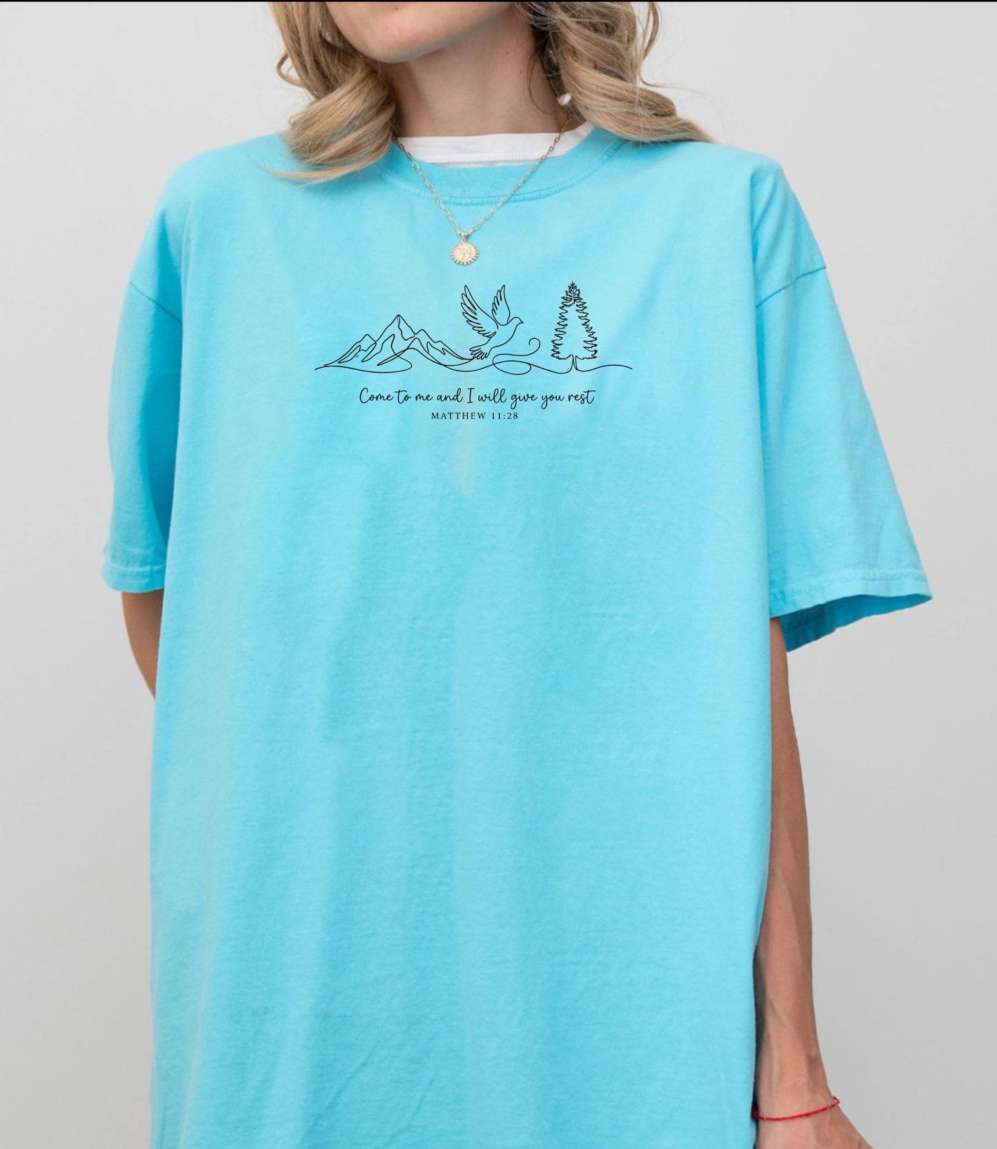 Person wearing a Lagoon blue t-shirt with nature-themed design and text that says "come to me and i will give you rest matthew 11:28" 