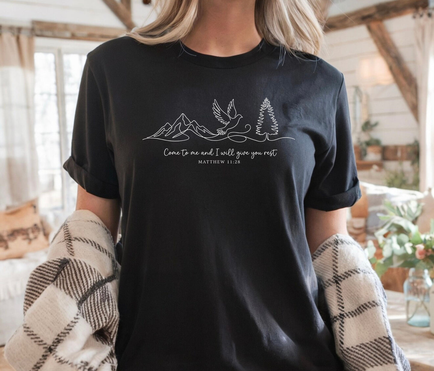 Person wearing a black t-shirt with nature-themed design and text that says "come to me and i will give you rest matthew 11:28" 