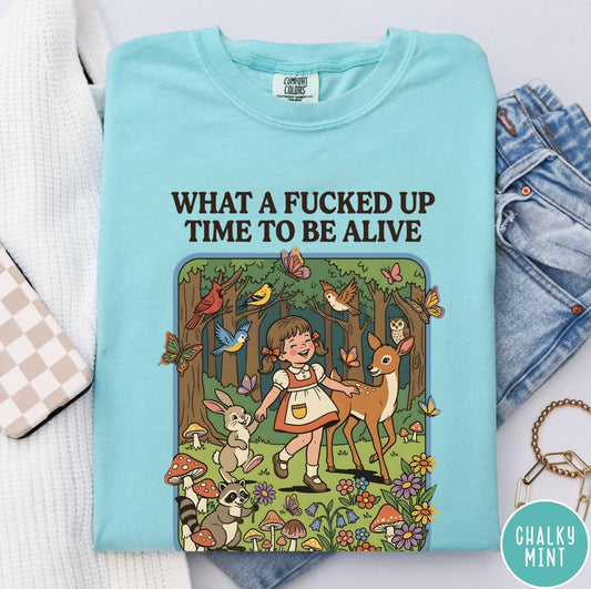 Light blue t-shirt with graphic design and text 'What a fucked up time to be alive' on a white surface.