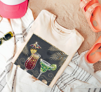 ivory t-shirt with colorful drink design on a sandy surface with accessories.