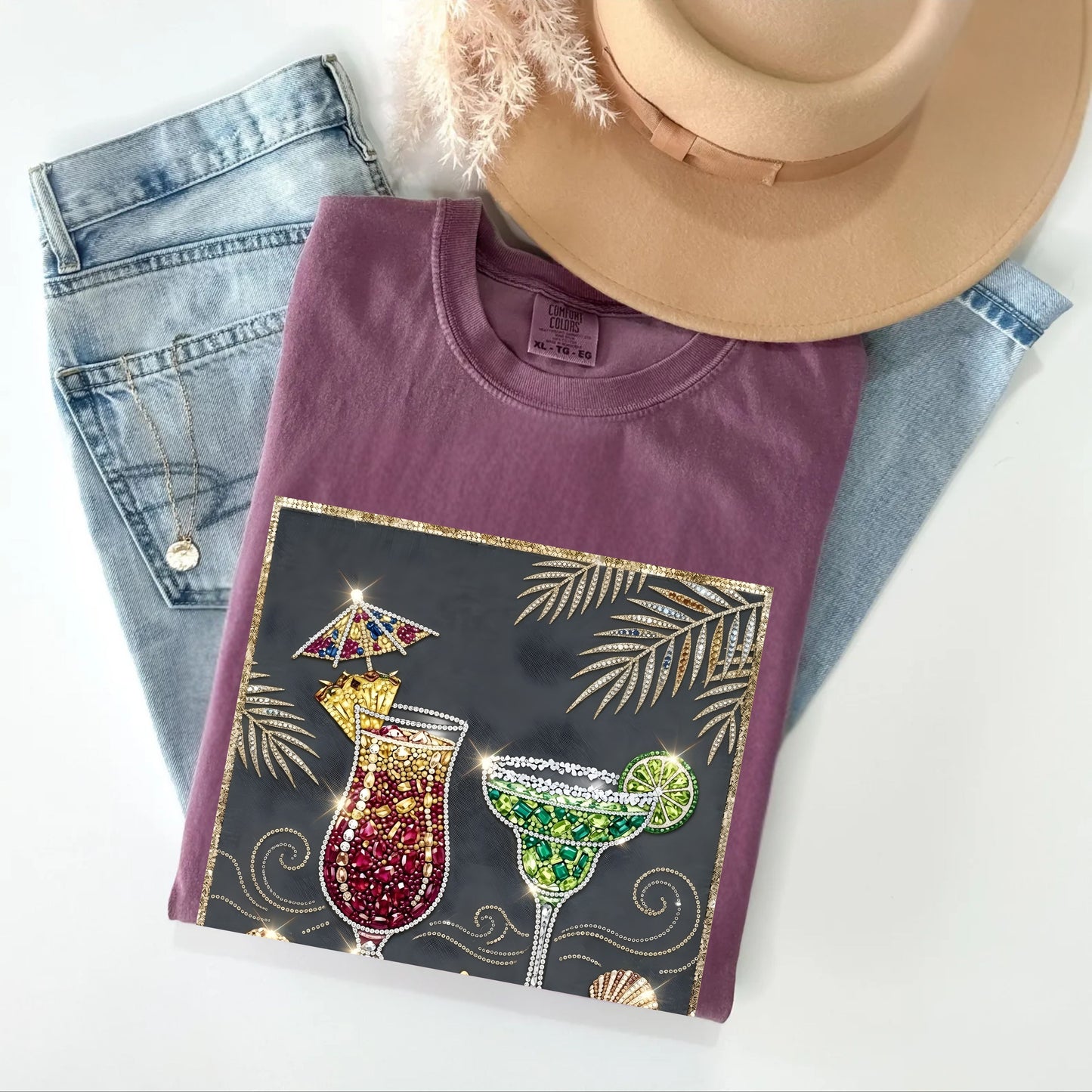 Folded berry t-shirt with cocktail design on a white surface with jeans and a hat.