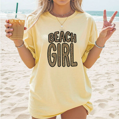 Person wearing a butter 'Beach Girl' shirt on a beach, holding a drink.