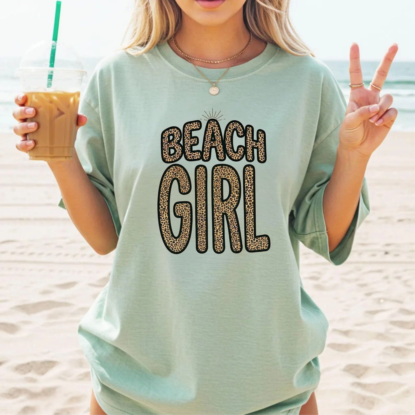 Person wearing a bay color 'Beach Girl' t-shirt on a beach, holding a drink.