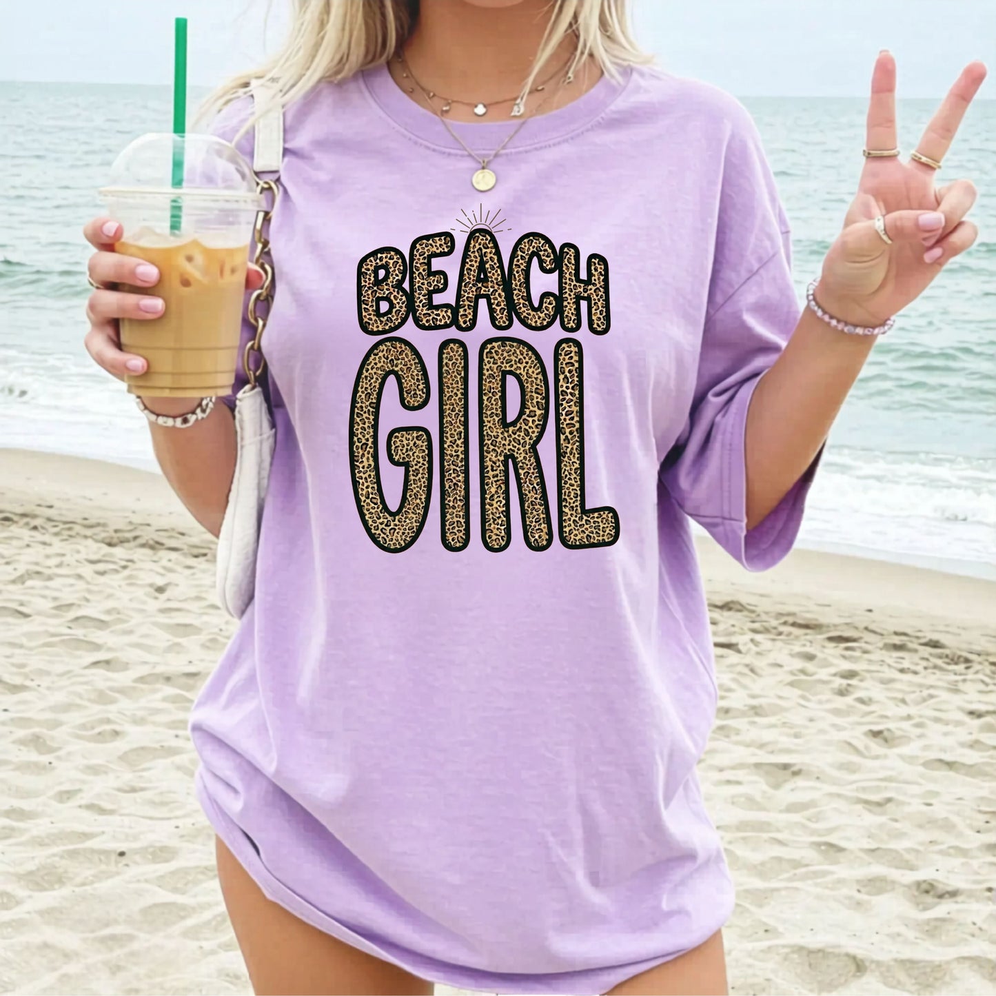 Woman on a  beach wearing a 'Beach Girl' orchid colored shirt, holding an iced coffee.
