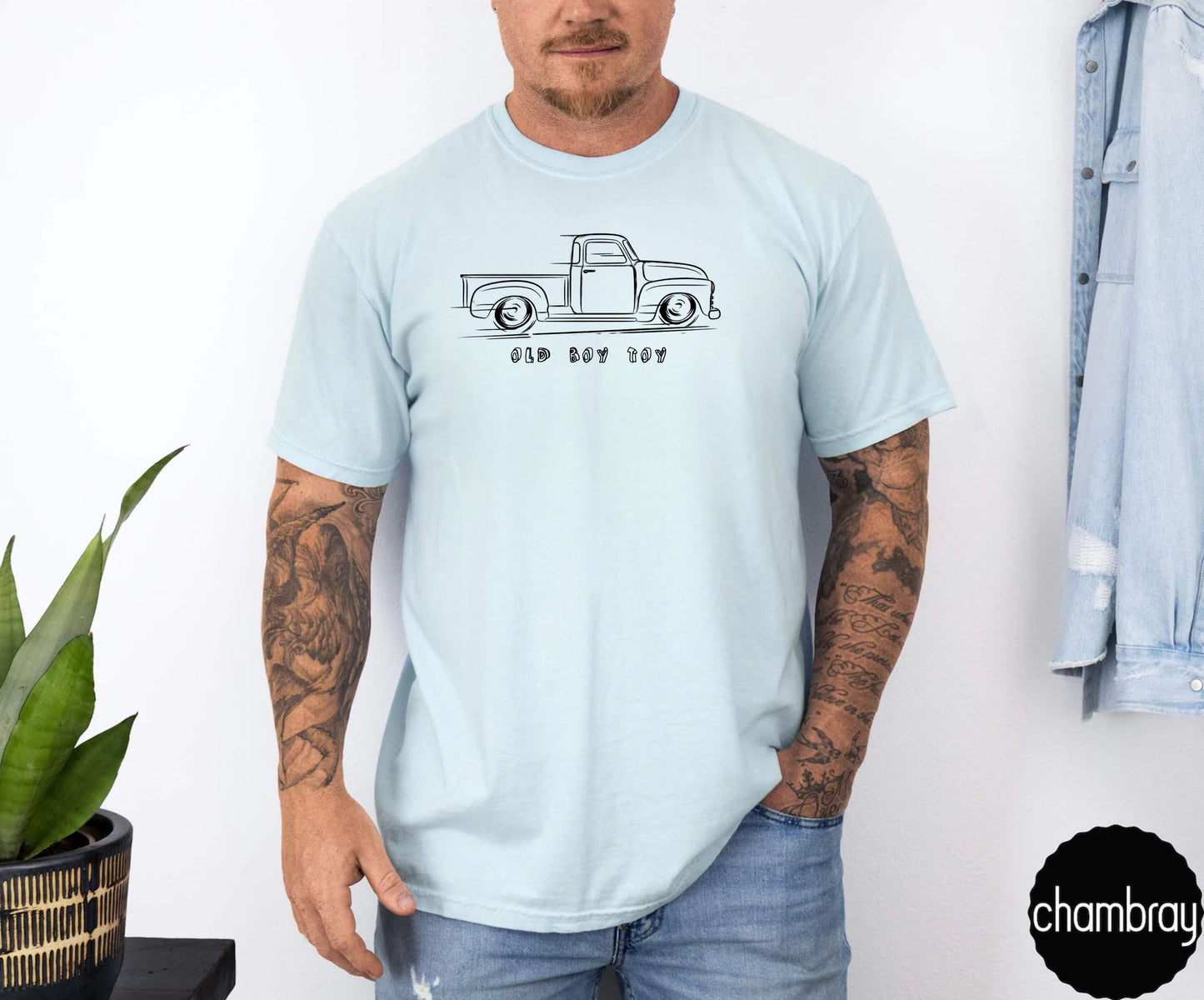 Person wearing a light blue t-shirt with a graphic design of old truck , standing against a white wall.