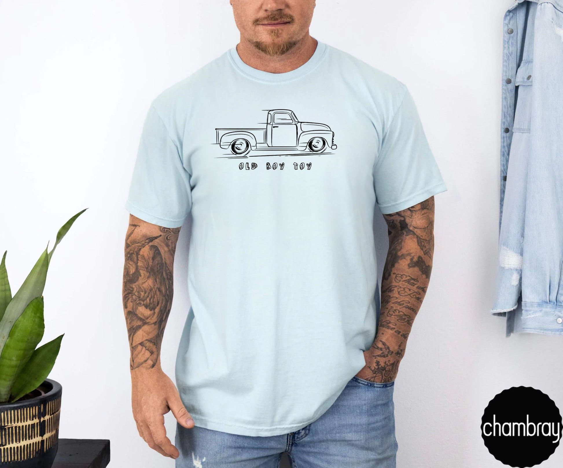 Person wearing a light blue t-shirt with a graphic design of old truck , standing against a white wall.