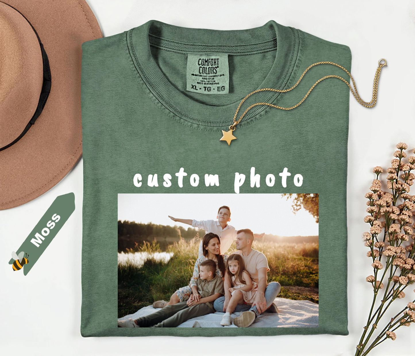 moss t-shirt with a family photo printed on it, placed on a white surface with a hat and dried flowers.